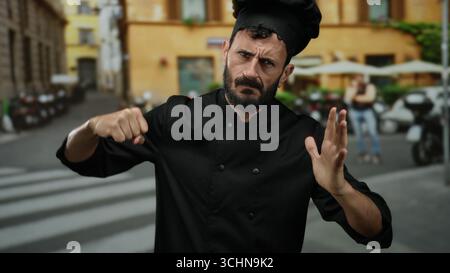 Homme hispanique barbu habillé comme chef imitant la pose de boxe dans une rue extérieure, mêlant tenue professionnelle et expression ludique. Banque D'Images