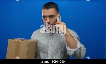 Jeune homme hispanique en chemise rayée tient une boîte d'expédition en carton collée et parle sur smartphone dans un studio bleu ; concentration. Banque D'Images