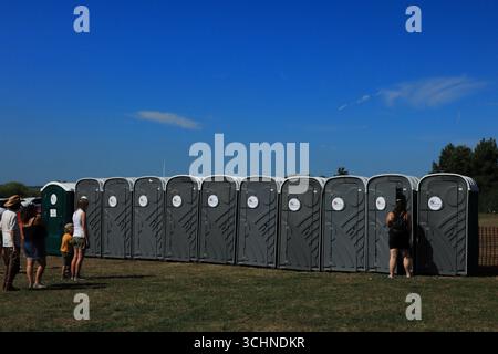 Une longue rangée de toilettes portables gris foncé est alignée sur un champ herbeux sous un ciel bleu clair. Quelques personnes sont debout à proximité, attendant de les utiliser ou de discuter. Au loin, d’autres structures d’événements et quelques arbres sont visibles. Illustration, éditorial : le rallye automobile de Stokes Bay à Gosport, Hampshire, Angleterre. 25 août 2025. Banque D'Images
