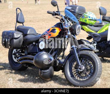 Illustration, éditorial : le rallye automobile de Stokes Bay à Gosport, Hampshire, Angleterre. 25 août 2025. Une moto Harley-Davidson noire avec des autocollants orange et jaune frappants est garée sur un terrain sec et herbeux. Il a un dossier, des sacoches en cuir noir, et des roues à rayons. Un casque noir repose sur le sol. Une autre moto avec un casque vert est visible derrière elle. Banque D'Images