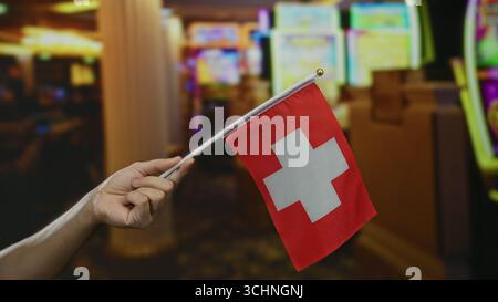 Homme tenant le drapeau suisse à l'intérieur du casino avec des machines à sous, représentant la fierté suisse dans le cadre récréatif. Banque D'Images