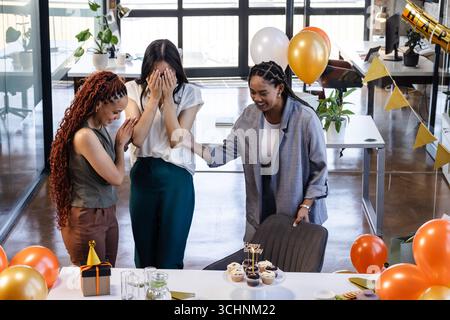 Collègues féminins divers célébrant une fête surprise au bureau avec des ballons et des cupcakes Banque D'Images