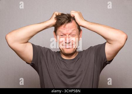 Homme très frustré avec des expressions faciales tirant les cheveux dans la photo de studio avec fond gris, homme d'âge moyen frustré européen, espace de copie Banque D'Images