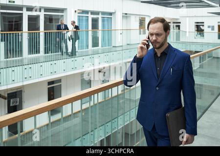 Jeune homme d'affaires caucasien marchant dans le couloir moderne de l'immeuble de bureaux parlant sur smartphone, tenant un étui d'ordinateur portable, deux cadres supérieurs masculins debout en arrière-plan conversant Banque D'Images