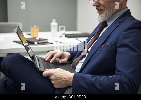 Homme caucasien d'âge moyen assis au bureau à l'aide d'un ordinateur portable dans un cadre de bureau, portant costume et cravate, se concentrant sur la frappe avec les mains sur le clavier, visage partiel visible Banque D'Images