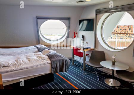Chambre sur DFDS Ferry Øresund Christianshavn Danemark // CHRISTIANSHAVN, Danemark — Une chambre à bord d'un ferry DFDS est présentée, avec un lit avec draps blancs et couvertures grises, un petit bureau avec chaise rouge et télévision, deux fenêtres circulaires offrant une vue sur l'eau et un bâtiment moderne en briques. La décoration de la chambre comprend de la moquette rayée et un papier peint texturé. DFDS est une compagnie maritime danoise qui exploite des liaisons de ferry dans toute l'Europe du Nord. Le ferry est amarré à Christianshavn, un quartier de canaux à Copenhague, au Danemark. Banque D'Images