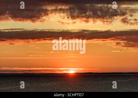 Lever de soleil sur la côte suédoise dans le Kattegat depuis Strömstad // Strömstad, Suède — Un lever de soleil vibrant illumine le ciel sur la mer du Kattegat, vu de la côte près de Strömstad dans le comté de Västra Götaland. Le soleil, un orbe lumineux à l'horizon, projette des teintes chaudes à travers les nuages et se reflète sur l'eau ondulée. La silhouette de la côte suédoise, avec des collines ondulantes et une tour de radio lointaine, forme une bande sombre contre le ciel dramatique. Ce spectacle naturel capture la beauté sereine de la côte scandinave au début d’une nouvelle journée. Banque D'Images