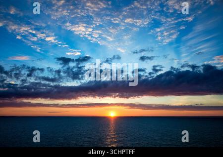 Lever de soleil sur la mer de Kattegat depuis Strömstad, Suède | STRÖMSTAD, Suède — Des nuages en couches bleues, orange et violettes encadrent le soleil levant au-dessus du Kattegat au large des côtes de Strömstad, dans le comté de Västra Götaland en Suède. Le Kattegat est le détroit qui sépare le Danemark et la Suède et relie la mer Baltique à la mer du Nord. Cette côte s'étend le long de la route des ferries de nuit reliant Copenhague et Oslo. Banque D'Images