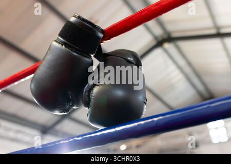 Les gants de boxe noirs pendent de la corde rouge au-dessus de la corde bleue sous les poutres brillantes du plafond Banque D'Images