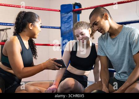 Divers amis de gymnastique s'appuyant sur le banc dans l'anneau de boxe vérifiant l'écran de smartphone avec des enveloppements de main roses Banque D'Images