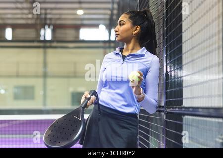 Femme indienne asiatique appuyée sur une clôture en maille sur un court de padel avec raquette, balle et smartwatch Banque D'Images