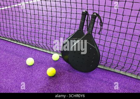 Deux raquettes de padel noir appuyées contre le filet sur un gazon violet avec des boules jaunes dispersées Banque D'Images