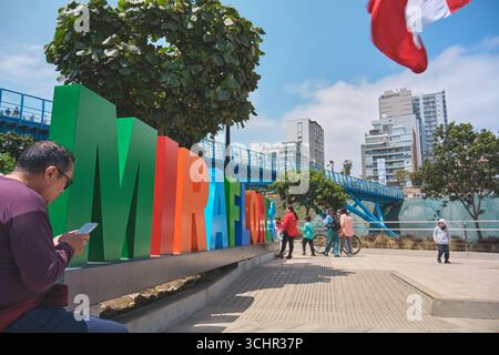 Lima pérou août 30 2025 : touriste utilisant un téléphone portable à côté du panneau coloré miraflores avec le nouveau pont la paz en arrière-plan Banque D'Images