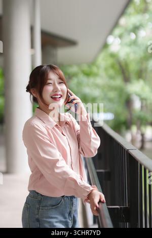 La femme adulte asiatique souriante se connecte via son smartphone elle trouve de la joie dans un environnement extérieur tout en mettant en réseau ou en gérant des tâches à distance Banque D'Images