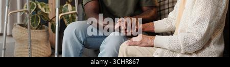 Jeune homme noir tenant la main avec une femme caucasienne senior assise ensemble à l'intérieur, montrant soutien et compassion, vue partielle avec des aides à la marche et plante en arrière-plan Banque D'Images