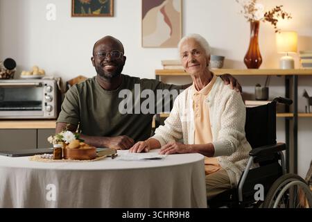 Portrait d'homme noir assis à côté d'une femme caucasienne senior handicapée en fauteuil roulant, souriant tous deux à la caméra, l'homme plaçant la main sur l'épaule de la femme, table avec des documents au premier plan Banque D'Images
