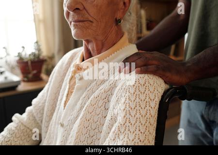 Femme caucasienne senior assise en fauteuil roulant avec un handicap, homme noir debout derrière plaçant doucement la main sur son épaule, les deux partiellement visibles dans un cadre intérieur Banque D'Images