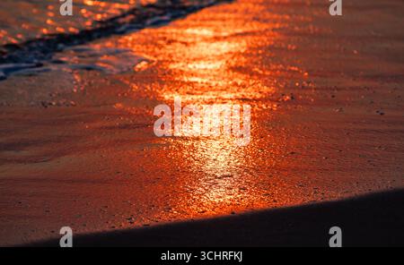 Un coucher de soleil serein se reflétant brillamment sur un rivage sablonneux à marée basse, mettant en valeur des teintes orange et rouge vives ainsi que de douces ondulations de l'eau, captu Banque D'Images