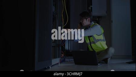Technicien portant gilet de sécurité, lunettes de connexion câbles dans la salle du serveur, avec smartphone, ordinateur portable Banque D'Images