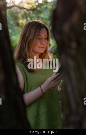 Une femme dans une blouse en lin vert lit un livre vintage à l'extérieur, encadré par des arbres dans une douce lumière du soir Banque D'Images