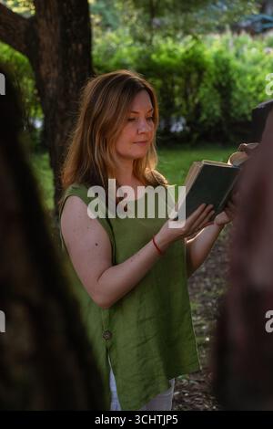 Une femme dans une blouse en lin vert lit un livre vintage à l'extérieur, encadré par des arbres dans une douce lumière du soir Banque D'Images