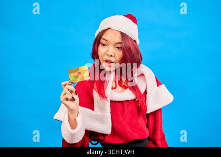 Jeune femme portant le costume de père noël regardant surpris tout en montrant la carte de crédit, sur fond bleu Banque D'Images