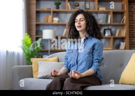 Jeune femme pratiquant la pleine conscience. Trouver la paix intérieure et profiter de la tranquillité tout en méditant sur un canapé confortable dans son moderne. Salon élégant. Créer une atmosphère apaisante et relaxante Banque D'Images