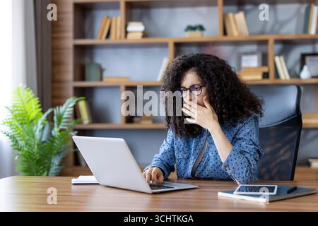 Entrepreneur féminin stressé couvrant sa bouche avec la main tout en lisant des nouvelles choquantes sur ordinateur portable, assis à un bureau en bois dans le bureau à domicile avec des étagères en arrière-plan Banque D'Images