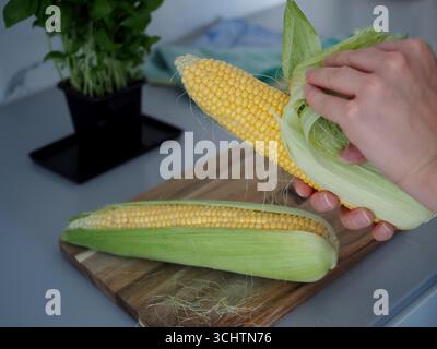 Mains préparant le maïs frais sur la planche à découper dans la cuisine Banque D'Images