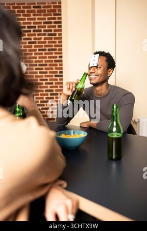 Homme afro-américain souriant assis à table et tenant une bouteille de bière, traînant avec des amis à la maison. Individu masculin noir avec carte sur le front, jouant au jeu de devinettes dans la salle de mur de briques. Banque D'Images