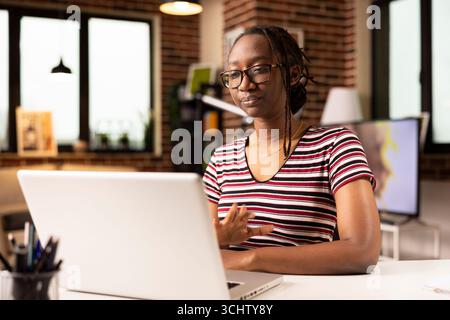 Entrepreneur afro-américain avec des lunettes assis à son bureau, s'engageant dans une session de formation virtuelle avec des collègues sur ordinateur portable. Femme d'affaires à distance participe à un webinaire professionnel à partir du bureau à domicile. Banque D'Images