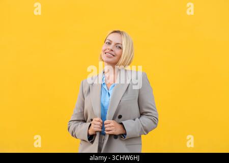 Jeune femme d'affaires avec les cheveux blonds courts portant blazer beige tenant blazer tout en souriant avec confiance sur fond jaune vif Banque D'Images