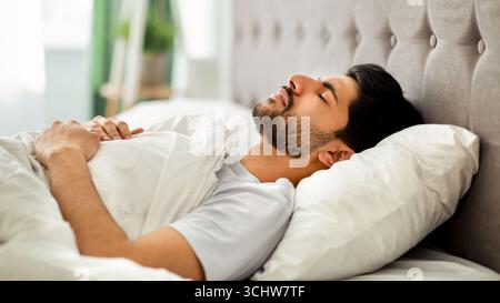 Sommeil profond chez les hommes. Un jeune homme arabe dormant paisiblement dans son lit confortable à la maison, allongé avec les yeux fermés sur le dos Banque D'Images