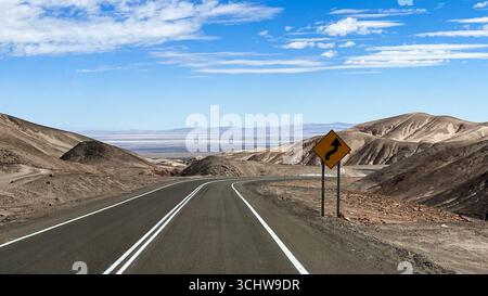 Route pavée avec panneau de signalisation jaune d'avertissement en plein air de jour contre le vent, tandis que la route tourne et passe à travers le paysage désertique à Atacama. Banque D'Images