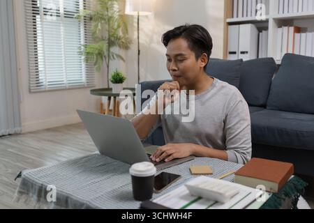 Un jeune homme est assis par terre à la maison, travaillant sur son ordinateur portable et pensant sérieusement. Il se concentre sur ses tâches en ligne, étudie ou travaille à distance Banque D'Images