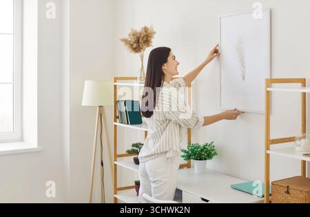 Femme joyeuse arrangeant une œuvre d'art sur le mur dans son appartement ensoleillé Banque D'Images