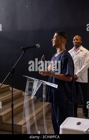 Homme dans les gommages détenant le prix, parlant au podium, collègue souriant en arrière-plan Banque D'Images