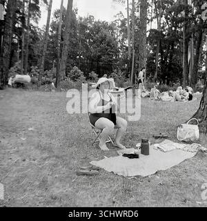 Une photographie d'archives en noir et blanc prise en URSS vers 1965. Une femme âgée en maillot de bain et un chapeau de soleil est assise sur une chaise pliante dans une forêt de pins et tricote lors d'une récréation en plein air. En arrière-plan, d'autres vacanciers prennent un bain de soleil dans une clairière. Le cadre transmet l'atmosphère authentique des loisirs soviétiques simples, un pique-nique d'été, ou le tourisme « sauvage » (dikari) dans les années 1960 Banque D'Images