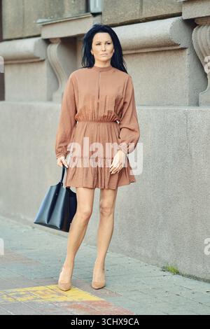 Une femme marche en toute confiance le long d'un trottoir occupé, portant une robe à la mode et portant un sac fourre-tout. Ses longs cheveux coulent doucement dans la brise comme elle Banque D'Images