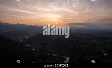 Vue aérienne de la lumière du soleil filtrant à travers les nuages sur des collines ondulantes et une rivière sinueuse, projetant de longues ombres à travers le paysage, Soppeng, Sulawesi Selatan, Indonésie. Banque D'Images