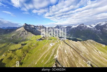 Vue aérienne de routes sinueuses traversant des montagnes verdoyantes et rocheuses, avec des sommets enneigés percant le ciel bleu vibrant, Bruck an der Großglocknerstraße, Salzbourg, Autriche. Banque D'Images