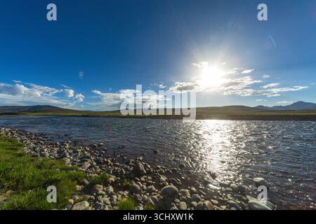 Vue de la lueur rayonnante du soleil se reflétant sur la surface de la rivière, encadrée par une rive rocheuse et des montagnes lointaines sous un ciel bleu vif, Deosai Nation Banque D'Images