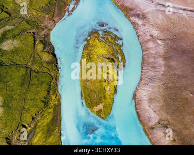 Vue aérienne d'une rivière turquoise vibrante sculptant à travers le paysage islandais, embrassant une île de mousse dorée sur fond de terr accidenté Banque D'Images