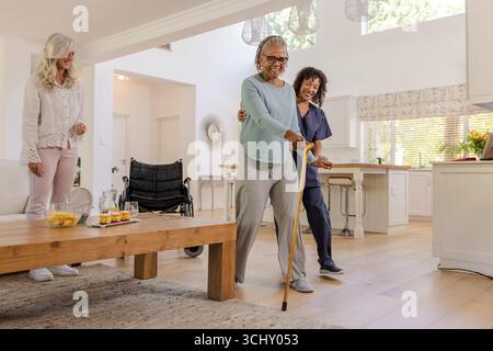 Femme âgée utilisant la canne de marche avec le soignant de soutien dans le salon lumineux Banque D'Images