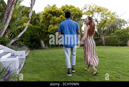 Jeune couple diversifié marchant main dans la main dans un jardin luxuriant, profitant d'une journée ensoleillée Banque D'Images