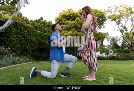 Couple diversifié, jeune homme proposant à la femme surprise dans le jardin, les deux regardant joyeux Banque D'Images