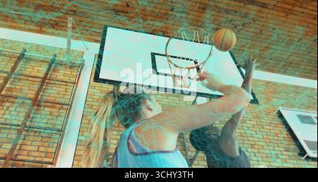 Atteindre l'athlète en maillot bleu tirant au basket-ball orange à la salle de gym, avec un adversaire masculin bloquant le cerceau Banque D'Images