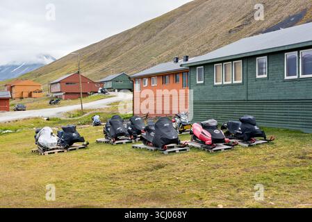 Motoneiges ski Doo Lynx Yamaha Longyearbyen Svalbard Norvège // LONGYEARBYEN, SVALBARD — plusieurs motoneiges, dont les modèles ski-Doo Lynx et Yamaha, sont stationnées sur des palettes en bois dans une zone herbeuse devant des bâtiments à Longyearbyen, Svalbard, Norvège. Les véhicules sont recouverts de bâches de protection, ce qui suggère qu'ils sont entreposés ou en attente de transport. Longyearbyen est la plus grande colonie et le centre administratif du Svalbard, un archipel situé entre l'océan Arctique et la mer du Groenland. Le paysage présente un flanc de montagne escarpé et rocheux en arrière-plan, typique de l'Arctique Banque D'Images