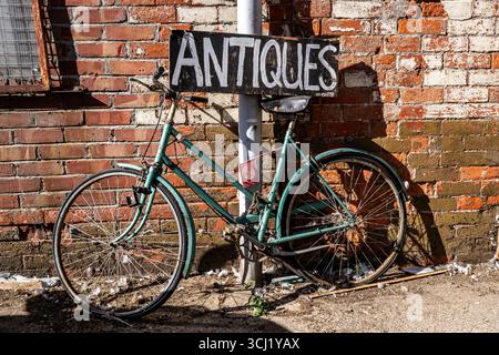 Une bicyclette vintage altérée repose contre un mur de briques rustique, sous un panneau « ANTIQUITÉS », capturé dans une nature morte détaillée. Banque D'Images