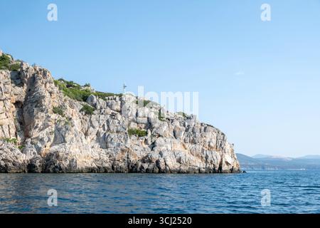 Falaise rocheuse de bord de mer avec un petit phare s'élevant au-dessus des eaux bleues près de Tolo, ciel bleu et montagnes lointaines Banque D'Images